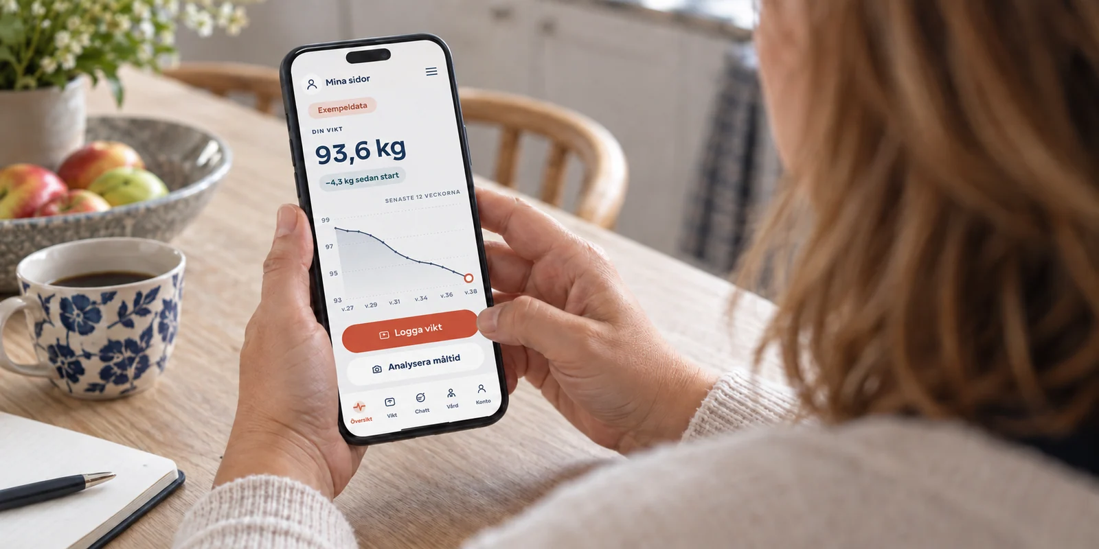 Över axeln på en kvinna som följer sin viktkurva på telefonen. Fiktiva exempeldata.