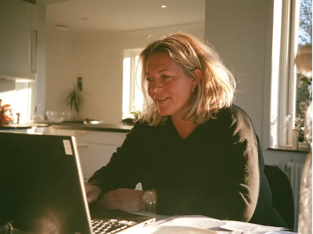 En kvinna sitter vid sin laptop i ett soligt kök och pratar med någon